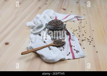 Vaso di vetro pieno di piccole lenticchie non cotte, cucchiaio di legno sul lato pieno di lenticchie e versato sul tavolo di legno, sulla parte superiore di un panno bianco da cucina. E Foto Stock