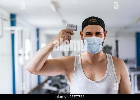 Uomo con maschera che prende la sua temperatura. Termometro a infrarossi. Palestra sana. Pandemia, coronavirus. Concetto di salute e sicurezza Foto Stock