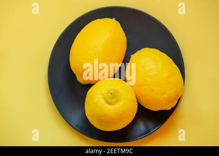 Tre limoni su un piatto nero su sfondo giallo. Background. Succo di agrumi, Foto Stock