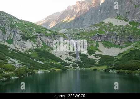 Riflessi nelle acque calme del lago, cascata e montagne al tramonto. Foto Stock