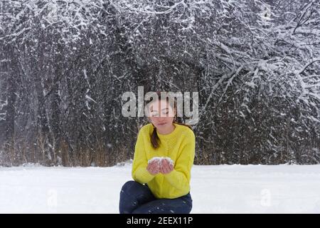 Giovane bella donna in inverno in giallo brillante maglione tiene la neve in mano e la guarda. Signora nella foresta innevata per una passeggiata. Foto Stock