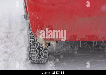 Ruota per auto con punte su strada innevata in inverno. Iciclette sul paraurti auto rossa. Concetto di guida pericolosa in condizioni di freddo estremo e maltempo. Foto Stock