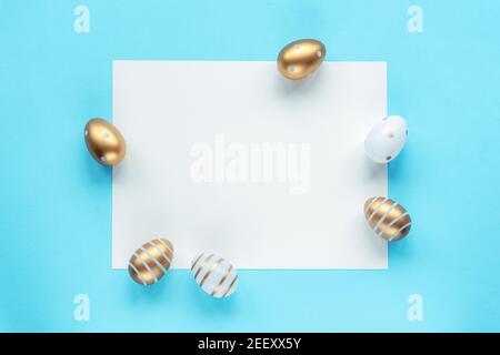 Uova di Pasqua dipinte in oro e bianco e foglio bianco su sfondo blu. Vista dall'alto, simulata. Foto Stock