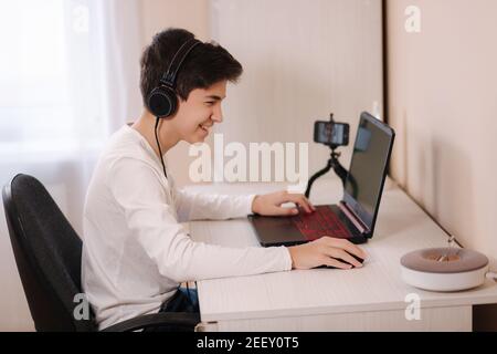 Teenage boy che gioca sul suo pc in una stanza bianca. I gamer acquisiscono video su webcam su laptop e telefono su treppiede Foto Stock