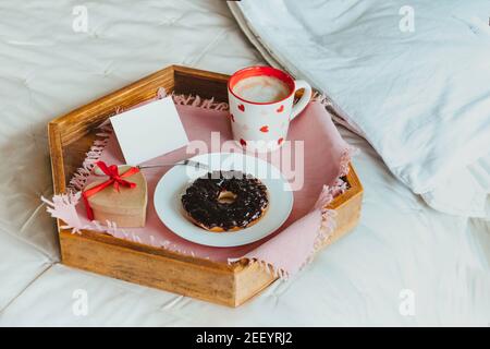 Semplice colazione di San Valentino a letto per l'amante. Una tazza di caffè e bagel con marmellata, scatola regalo a forma di cuore con nastro rosso, carta vuota vuota per Foto Stock