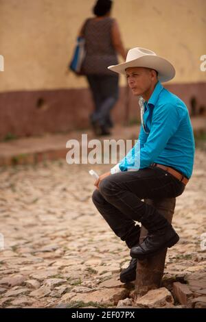 Un giovane in cappello da cowboy , camicia blu brillante e stivali neri si siede in attesa sulle strade di Trinidad a Cuba Foto Stock
