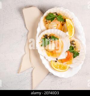 Capesante al forno con caviale e salsa cremosa all'aglio in piatto bianco. Capesante con limone su fondo bianco con tovagliolo. Vista dall'alto. Spazio di copia. Disposizione piatta Foto Stock