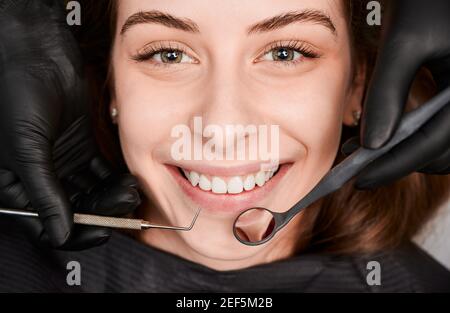 Primo piano di bella giovane donna che guarda la macchina fotografica e sorridente mentre dentista tiene gli strumenti dentali. Le mani del dentista che usano l'esploratore dentistico e lo specchio mentre esaminano i denti della paziente femminile. Foto Stock