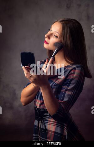 Ragazza polveri il suo viso con un pennello guardando nello specchio, studio foto su uno sfondo grigio Foto Stock