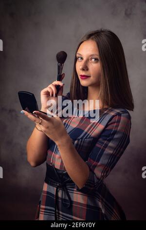 La ragazza si spolvero il viso con un pennello mentre si guarda nello specchio, la ragazza è stata distratta e guardata nella cornice, studio di fotografia su una ba grigia Foto Stock