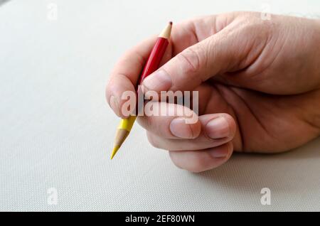 Un uomo a destra tiene una matita colorata affilata su entrambi i lati su un tavolo bianco. Matita rossa e gialla nella mano dell'uomo adulto. Primo piano. Selettivo fo Foto Stock
