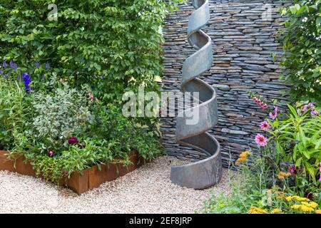 Piccolo giardino cottage cortile inglese con scultura a spirale - muro di pietra a secco - siepe - percorso di ghiaia con giardino fiorito confini Inghilterra UK Foto Stock