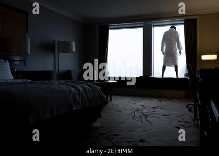 Uomo infelice che si trova nella finestra della camera d'albergo, guardando giù per la strada e pensando di suicidarsi. Figura maschile in accappatoio in retroilluminazione. Concetto di disperazione e depressione. Foto Stock