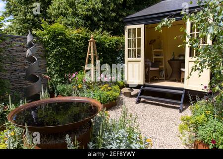 Un piccolo cottage all'inglese con Summerhouse Shepherds Hut in un giardino di ghiaia - lavoro da casa ufficio - acqua con scultura a spirale obelisco UK Foto Stock