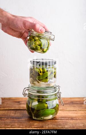 Mano maschile tenendo uno dei tre vasi di vetro di peperoni Jalapeno appena decapati con erbe e aglio. Copia spazio Foto Stock