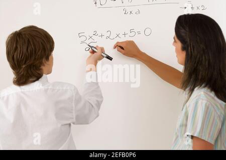 Vista posteriore di un ragazzo adolescente che risolve algebra su lavagna bianca e un'insegnante femminile che sta accanto a lui Foto Stock