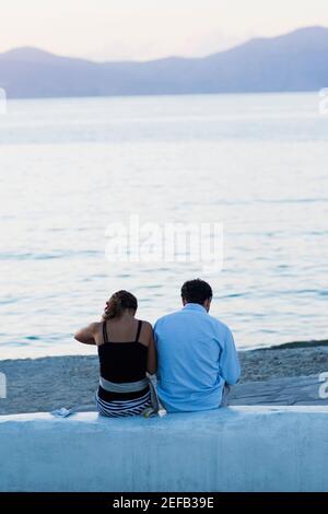 Vista posteriore di una coppia seduta su un muro vicino a una spiaggia, Mykonos, Cicladi, Grecia Foto Stock