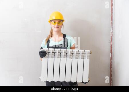 Donna idraulico tiene bianco centrale riscaldamento radiatore, in tute e cappello giallo contro parete grigia. Foto Stock