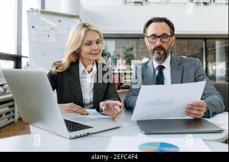 I partner aziendali maturi si concentrano imparando grafici finanziari per il nuovo progetto, facendo documenti cartacei. Donna elegante e uomo serio in abiti formali seduti alla scrivania, guardare i documenti, lavoro di squadra Foto Stock