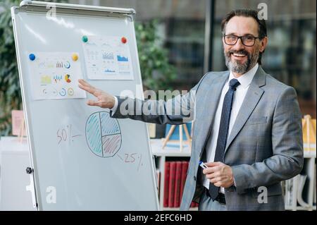 Un imprenditore maturo intelligente e sicuro fa una presentazione in conferenza, suggerisce una nuova strategia aziendale per l'azienda. Un impiegato maschile di successo in SUIT sta in piedi vicino alla lavagna a fogli mobili in ufficio, sorride a. Foto Stock