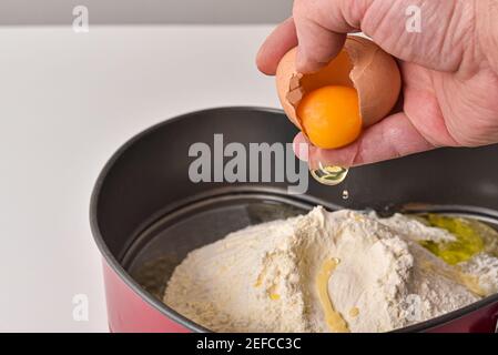 La mano maschile rompe l'uovo in farina mentre si fa l'impasto. Primo piano della mano dello chef con il tuorlo d'uovo giallo nel processo di preparazione per la cottura. Foto Stock