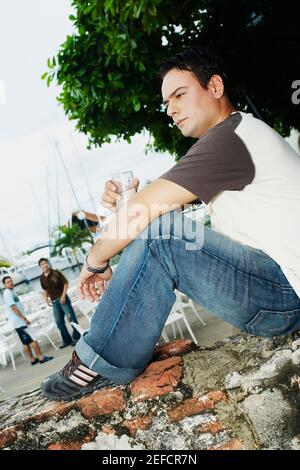 Profilo laterale di un giovane uomo seduto su un mattone muro e tenendo un bicchiere d'acqua Foto Stock