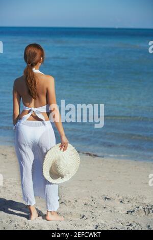 Vista posteriore di una donna in piedi sulla spiaggia e. tenendo un cappello di paglia Foto Stock