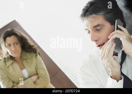 Uomo maturo che parla segretamente su un telefono cellulare con la moglie che lo guarda sospettosamente Foto Stock