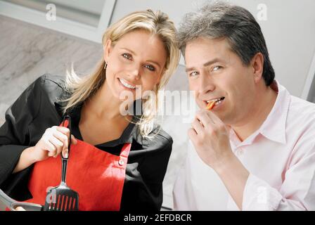 Primo piano di una donna di mezza età adulta che cucinava cibo con un uomo maturo che mangia cibo in cucina Foto Stock