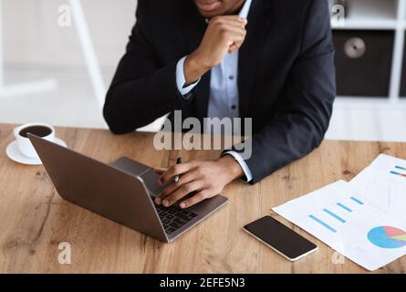 Troncato di uomo d'affari nero in tuta che lavora con il laptop in ufficio, controllo del rapporto di marketing, tecnologie moderne nel concetto di business. Irriconoscibile Foto Stock