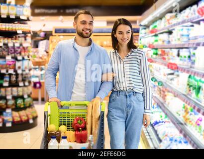 Happy Millennial Family Couple acquistare cibo in Supermarket, scegliendo prodotti Walking con carrello lungo le corsie e ripiani completi acquistare alimentari Toge Foto Stock