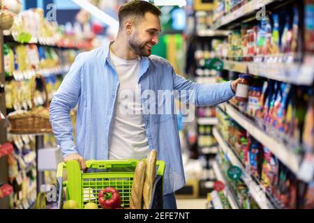 Concetto di shopping quotidiano. Sorridente giovane uomo che spinge carrello del trolley tra le corsie nel negozio di alimentari. Positivo Guy che acquista cibo, prendendo prodotti da Shel Foto Stock