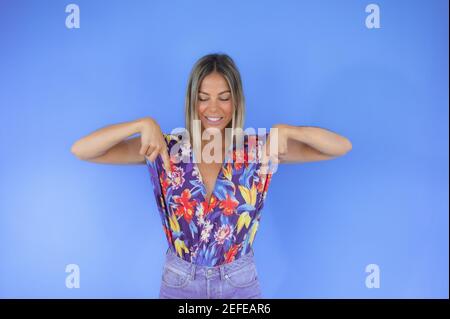 Donna in camicia floreale e jeans in piedi che punta a. terra con entrambe le mani su sfondo blu Foto Stock
