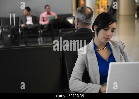 Donna d'affari che usa un laptop con un uomo d'affari dietro di lui a. un aeroporto Foto Stock