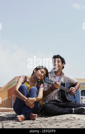Giovane uomo seduto con una giovane donna e di suonare una chitarra Foto Stock