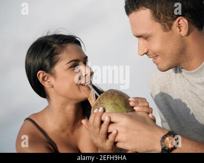 Primo piano di una giovane donna che beve acqua di cocco da un cocco tenuto da un giovane uomo Foto Stock