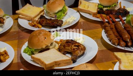 Piatti pronti al tavolo da cucina, la famiglia si siede insieme per la cena. Serve pollo al barbecue con hamburger e salsicce alla griglia con pane. Foto Stock