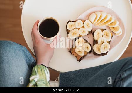 Donna tiene una tazza di caffè nero, sparato direttamente sopra. Avere frutta sana e panino burro di arachidi per la colazione, bere il caffè del mattino a casa Foto Stock
