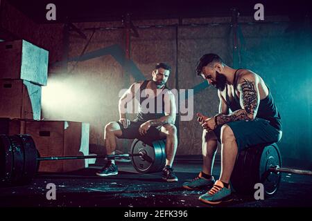 Due muscolare tattoed barbuto atleti rilassarsi dopo la formazione, il sollevamento di un carico pesante peso. fumo in palestra. Urlo. Lavorando sodo. Esercizi per i muscoli della schiena Foto Stock