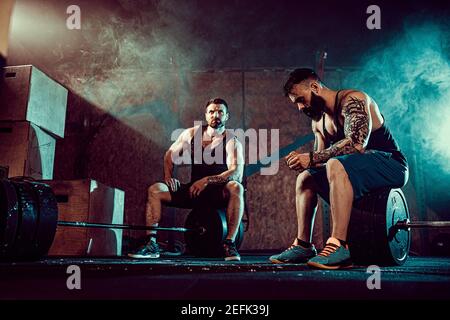 Due muscolare tattoed barbuto atleti rilassarsi dopo la formazione, il sollevamento di un carico pesante peso. fumo in palestra. Urlo. Lavorando sodo. Esercizi per i muscoli della schiena Foto Stock