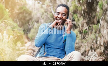 Giovane uomo di etnia africana che parla via cellulare con qualcuno e ridendo allegro mentre ha una passeggiata a piedi nella foresta tropicale. Felice Peopl Foto Stock