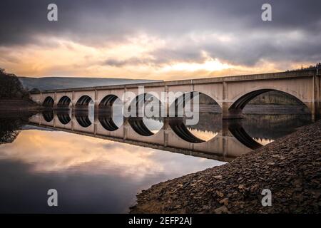 Lungo ponte ad arco che attraversa il lago artificiale di Ladybower riflesso nella valle di Derwent ancora acque profonde del fiume con alba bella cornice di cielo arancione dietro Foto Stock