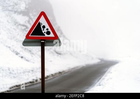 Cartello di avvertimento rosso triangolo che cade rocce su neve ghiacciata coperta tortuosa strada di montagna che passa attraverso fossi nebbia piena valle tra colline durante l'inverno Foto Stock