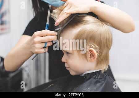 Parrucchiere taglia i capelli serio stretto bambino ragazzo biondo. Primo piano sullo stile di vita. Concetto di bellezza, igiene. Foto Stock