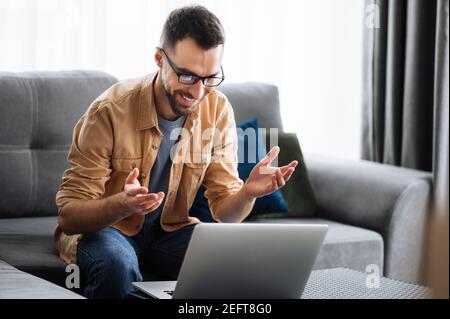 Freelance maschile intelligente che utilizza un computer portatile, videochiama, comunica con colleghi o amici tramite videoconferenza e sorridendo, lavorando da casa. Concetto di lavoro remoto Foto Stock