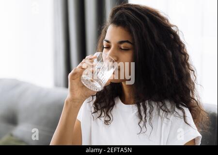 Stile di vita sano. Giovane donna afroamericana beve un bicchiere d'acqua mentre si siede sul divano a casa. Bella donna sana seguire uno stile di vita sano Foto Stock