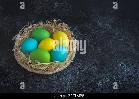 Uova di pasqua colorate in piastra di legno decorata con trucioli decorativi su sfondo scuro. Spazio di copia Foto Stock