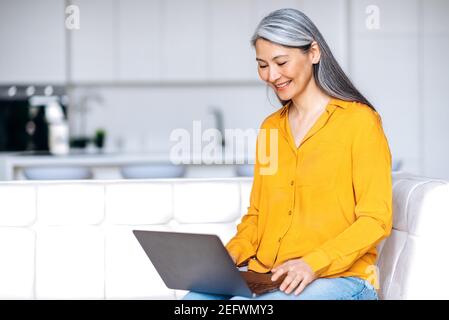 Rilassata donna asiatica con capelli grigi di mezza età vestita di abbigliamento casual elegante si siede a casa in salotto, utilizzando il laptop, acquistando online, lavorando da casa o studiando a distanza, navigare sul social network Foto Stock