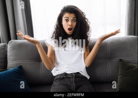 Ritratto di sorpresa afroamericana ragazza con capelli ricci. Shocked giovane donna in abbigliamento casual si siede sul divano a casa, ha ottenuto notizie inaspettate, si ritrae, e confuse guarda direttamente la fotocamera Foto Stock