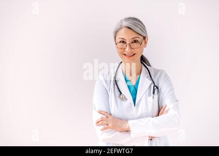 Ritratto di un sicuro medico asiatico con capelli grigi in uniforme medica e stetoscopio si trova su isolato sfondo bianco con le braccia incrociate, guarda la fotocamera e sorride Foto Stock
